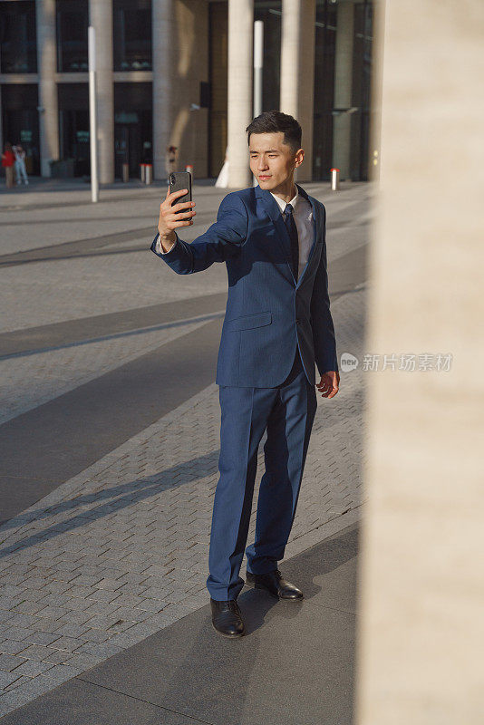 一名身穿蓝色经典西服白衬衫的年轻亚洲业务员在商业中心前的广场上自拍，照片的前景是广场柱子