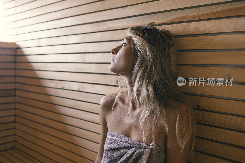 水疗日:美丽的年轻金发女子坐在桑拿浴室裹在灰色毛巾