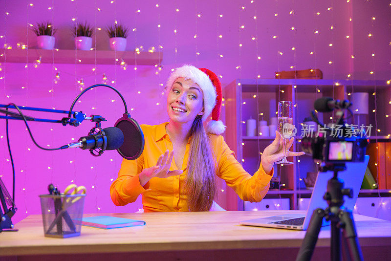 一个穿着黄色衬衫，戴着圣诞老人帽子的金发博主在紫色背景的工作室里为她的粉丝录制庆祝祝酒词