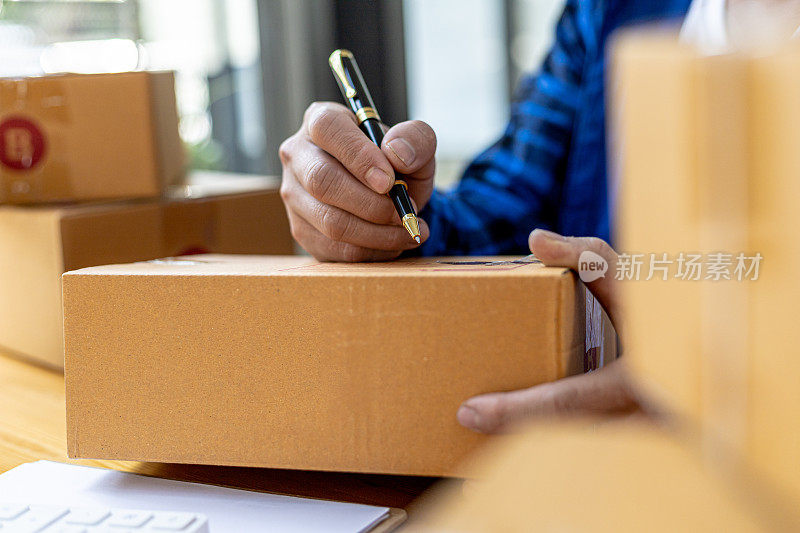 亚洲人写客户的运输信息在包裹盒子，他拥有一个网上商店，他通过一个私人快递公司运送产品给客户。在线销售的概念。