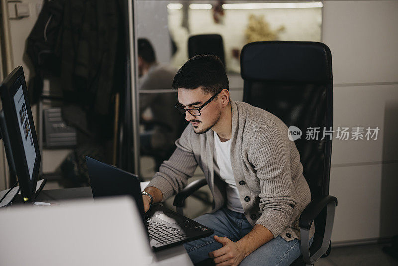 年轻人戴着眼镜，坐在家里的桌子上用笔记本电脑上网。