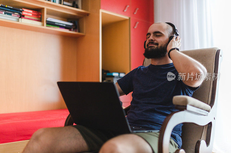 秃顶的年轻人在家里使用笔记本电脑，一边听着耳机里的音乐