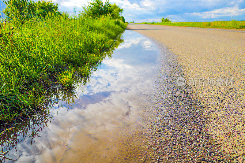 潮湿的柏油路，路上的水洼，天空的倒影和绿色的草地。
