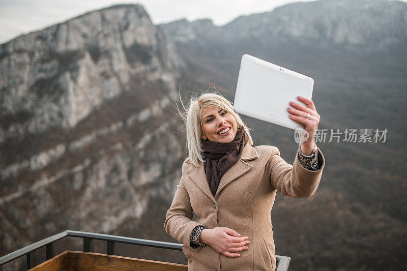 在寒冷的冬天，一个微笑的女人在山顶上使用电子平板电脑