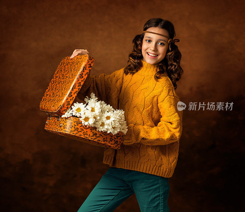 这个女孩手里拿着一个橘黄色的旧手提箱。她头上戴着一条缎带。手提箱里放着一束白色的菊花，雏菊。女孩微笑。照片在复古风格。