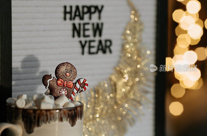 一个写着“新年快乐”的牌子，一个装有热可可、棉花糖的马克杯，一个姜饼人以美丽的黄色焦景为背景。一个温馨的圣诞和寒假概念。