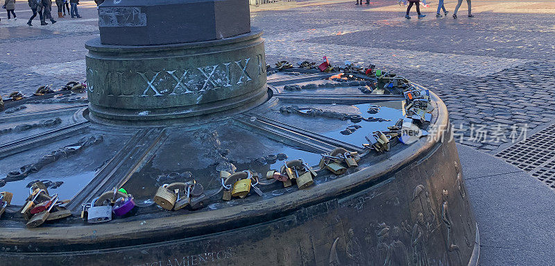 在一个广场灯柱的圆形底座上的爱情挂锁的特写。背景是不认识的人在广场上行走。