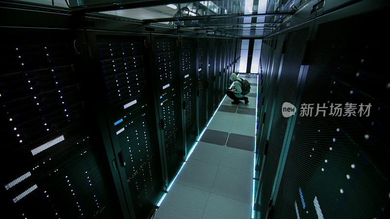 一个蒙面黑客在数据中心的安全摄像头视图，与他的笔记本电脑，他连接到机架服务器之一，犯罪。