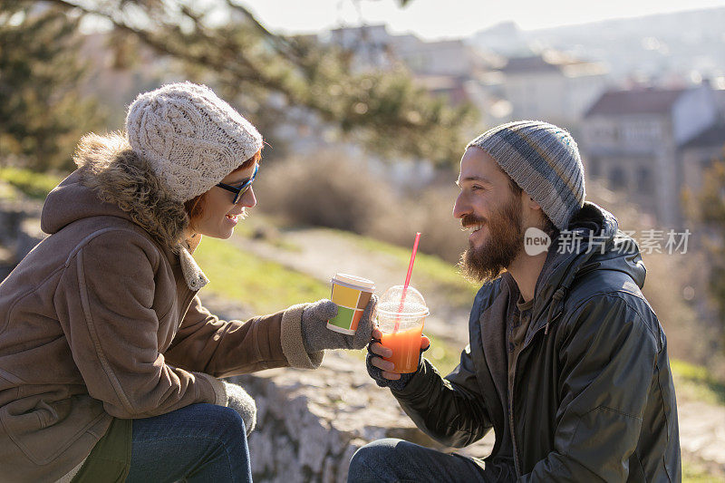 戴着冬季帽子的年轻夫妇坐在公园长椅上聊天喝饮料