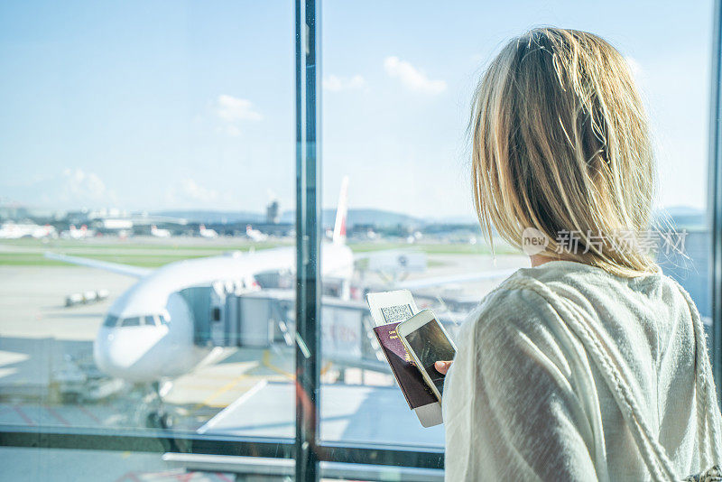 在机场等待航班的年轻女子坐在长凳上，手里拿着手机和护照