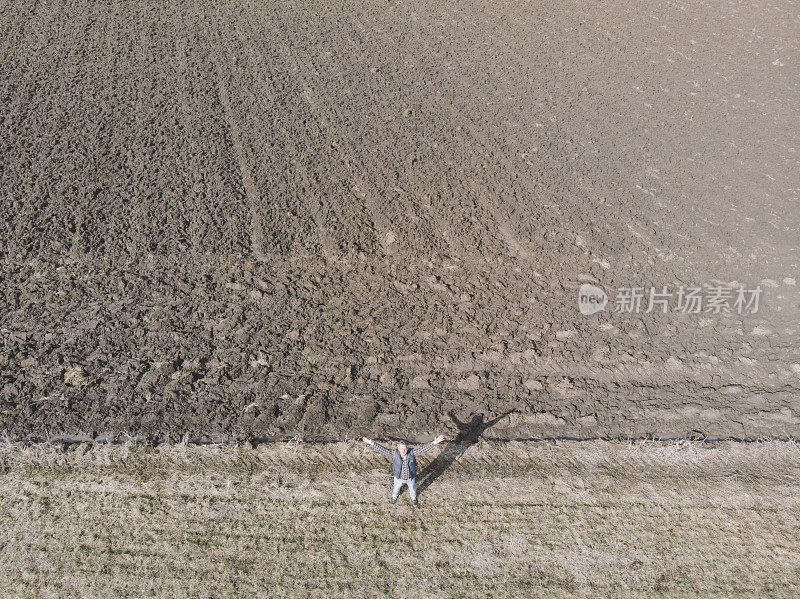 英俊的成年农民站在他的犁过的农田里，双臂伸开在冬末。鸟瞰图。