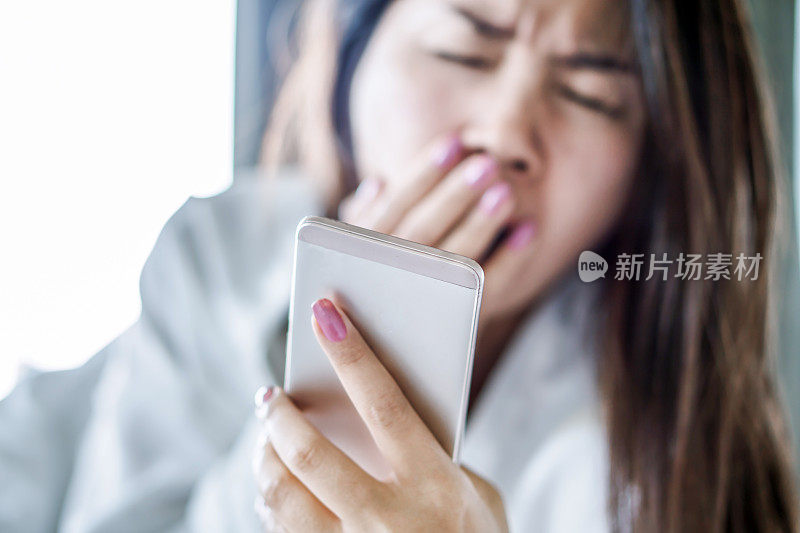 亚洲女人在床上打哈欠，手拿着智能手机感觉疲倦和困倦