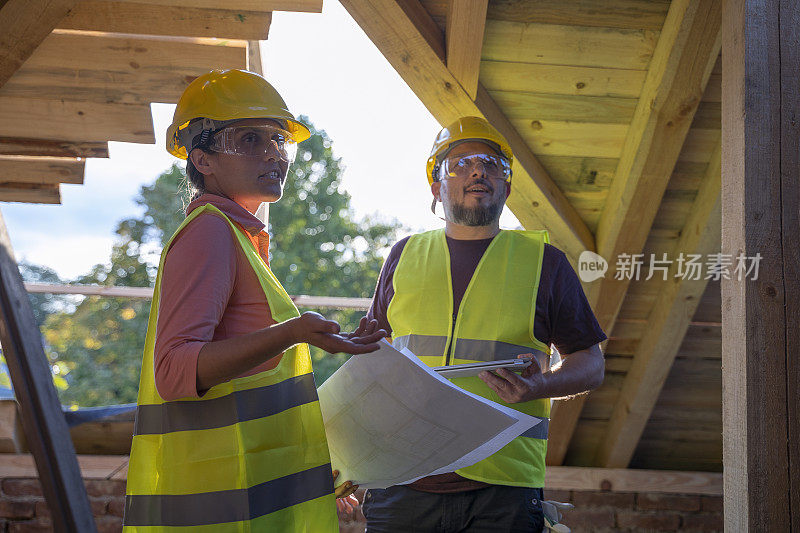 盖屋顶。一个阳光明媚的日子，一位自信的女建筑师和一位建筑工人在住宅楼的工地上一起讨论和工作。