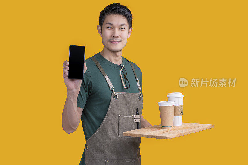 聪明友好的亚洲男性眼镜咖啡师小咖啡店老板在围裙的手势欢迎顾客来享受他的咖啡饮料，快乐的亚洲男性展示智能手机在线订购为您准备好