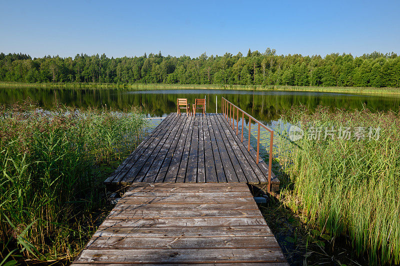 在阳光明媚的日子里，木制码头上的两把木椅俯瞰着平静的湖泊和森林。放松的概念，