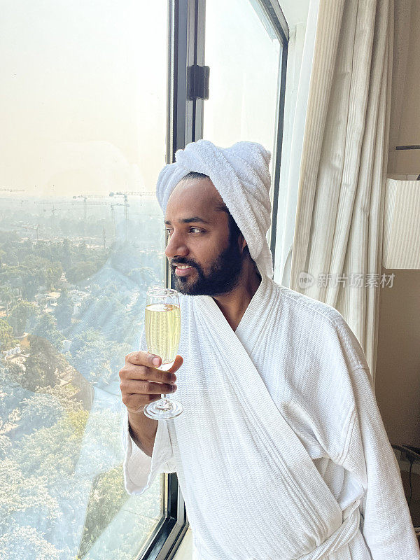 特写图像的印度男子站在酒店房间靠在窗户喝香槟从香槟笛子，穿着白色毛巾长袍和毛巾包裹头部，标准灯，焦点在前景