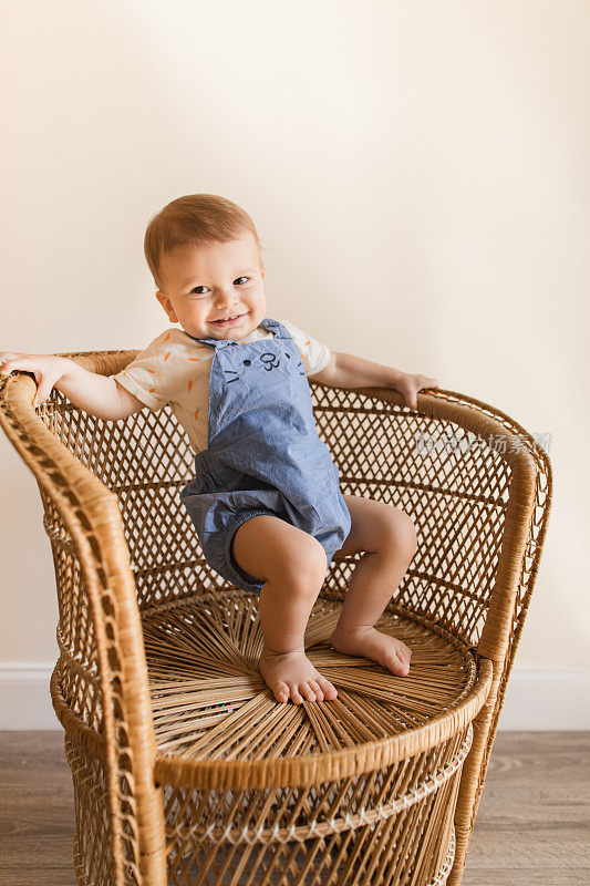 一个15个月大的古巴裔美国男婴，浅棕色短发和棕色眼睛，穿着胡萝卜连体衣和蓝色兔子工作服，还有蓬松的兔子尾巴，光着脚，淘气地站在复古藤椅上