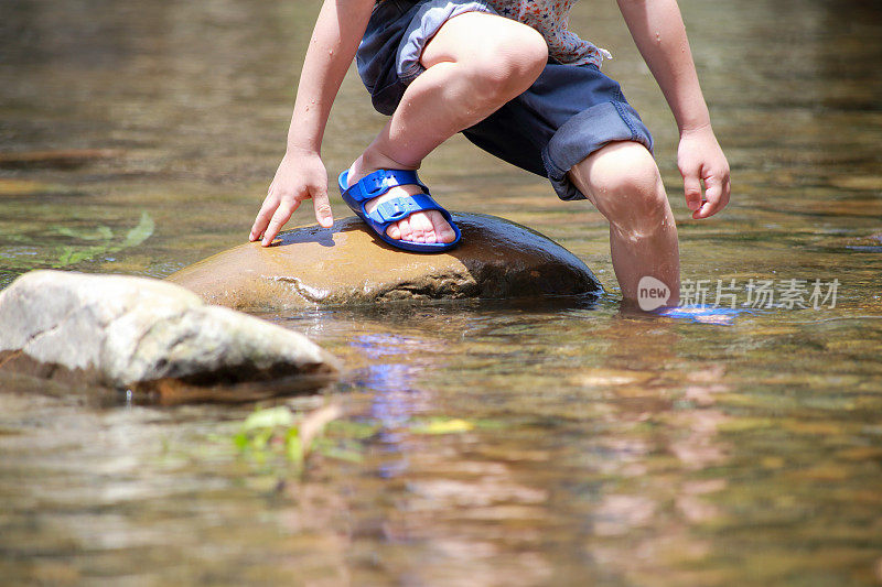 亚洲男婴穿着拖鞋站在小溪里的一块石头上
