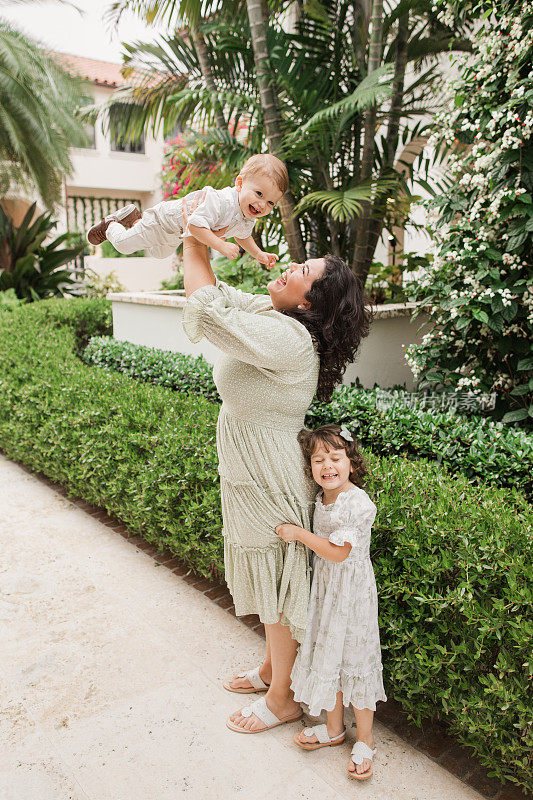 一位快乐的31岁母亲带着她4岁的古巴裔美国女孩和1岁的男婴穿着鼠尾草绿和白色的春装，在佛罗里达州棕榈滩的托斯卡纳风格的花园里