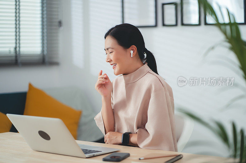 亚洲女性戴着耳机，喜欢坐在家里的办公室里，用笔记本电脑在线工作，进行视频会议。
