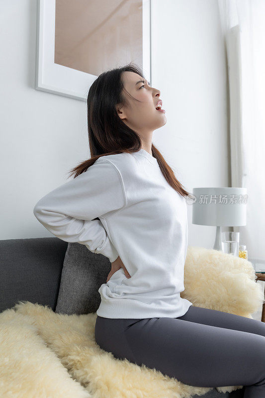 年轻漂亮的亚洲女孩坐在客厅舒适的沙发上，背部疼痛。
