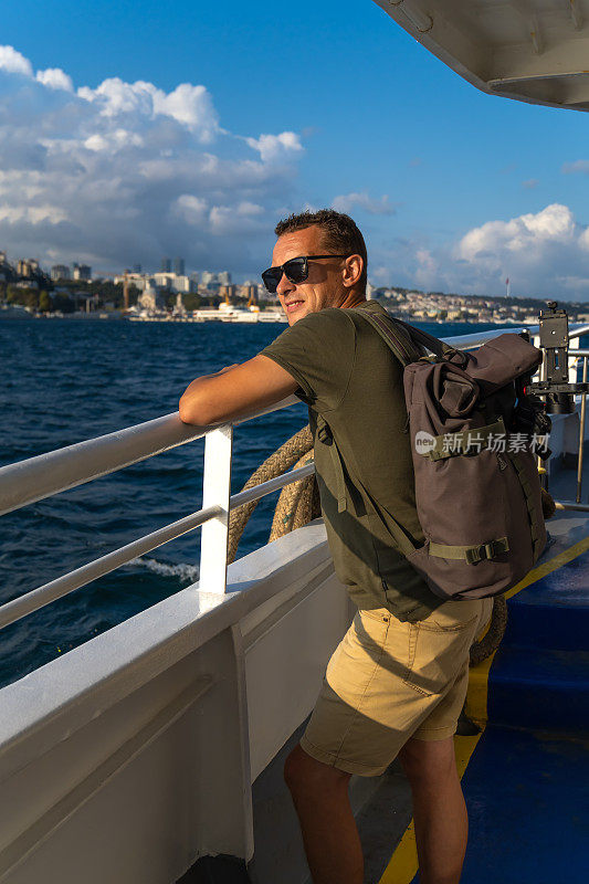在一个阳光明媚的日子里，快乐的年轻男游客背着背包在伊斯坦布尔的渡轮上享受着大海和这座城市。旅游的概念，旅游。垂直图