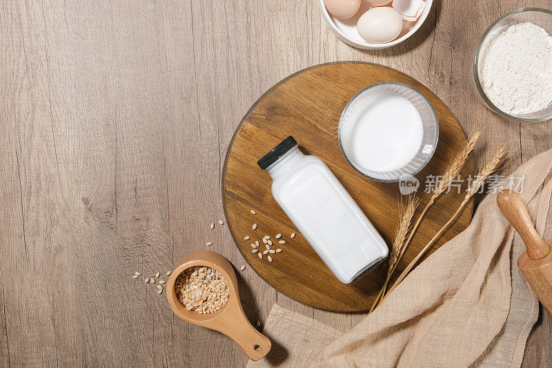 平躺在浅色的木制背景上，一个圆形的木制托盘里放着一瓶牛奶和一杯牛奶，上面装饰着面粉、鸡蛋、大麦等其他食材。复制空间，俯视图