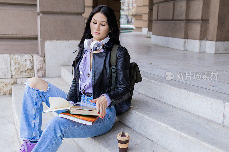 年轻漂亮的女大学生背着双肩包坐在大学楼梯上。她戴着耳机，穿着皮夹克，等待着大学课程的开始。