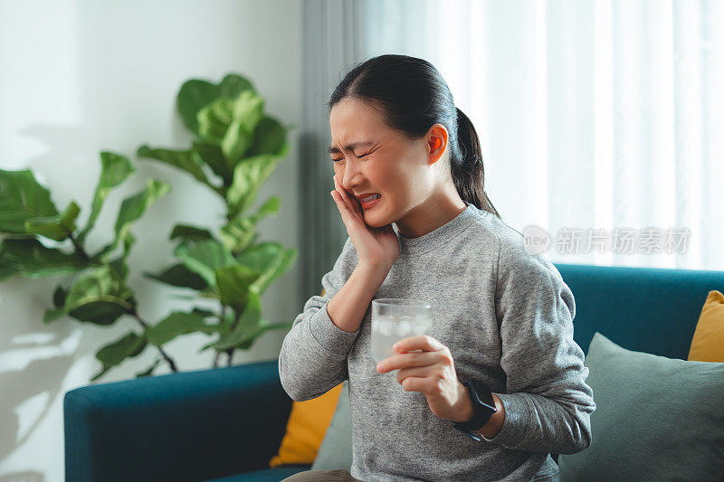 亚洲女性坐在客厅的沙发上，喝了凉水，摸了摸脸颊，就会感到牙齿敏感。