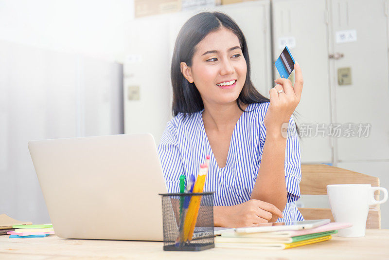 快乐的亚洲女人在网上购买笔记本电脑和支付信用卡在她的家庭办公室