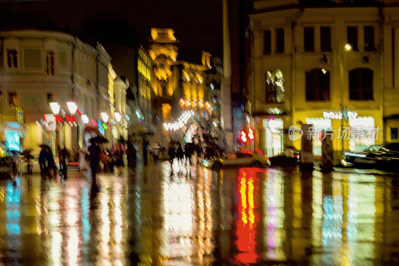 抽象的背景，不认识的人匆忙在城市街道在雨夜。商店橱窗里明亮的灯光，街灯，都模糊了。现代城市生活方式