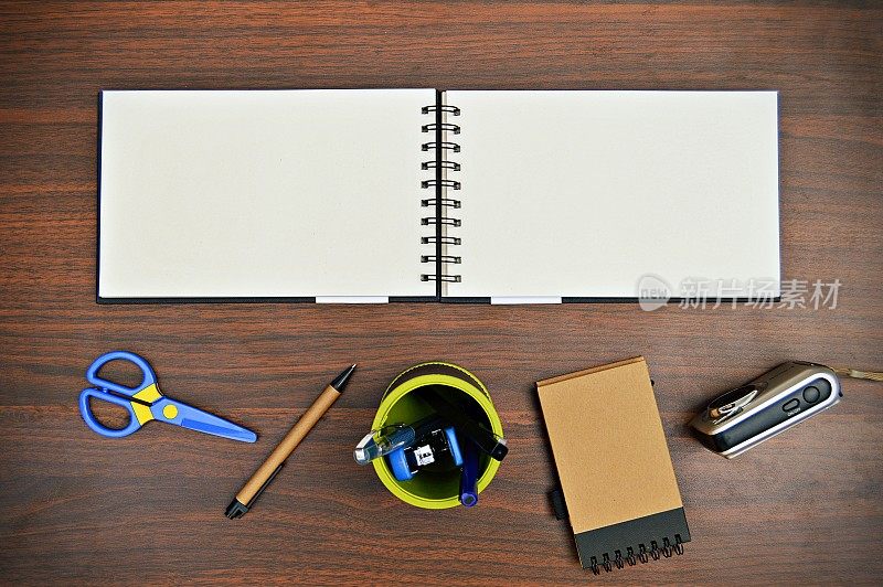 一幅文具的横向照片，如钢笔、日记本、剪刀、笔架、订书机、照相机，放置在一个木制的深棕色水平背景上，非常美观。