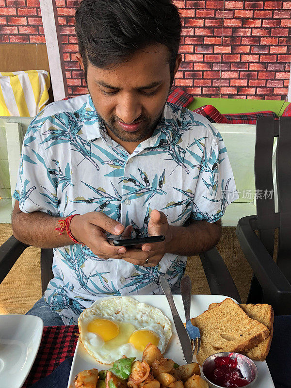 年轻男子看着手机，吃着传统的英式早餐，比如煎蛋、面包、土司，微笑着看手机短信，用手指滚动屏幕，查看社交媒体，网络新闻，穿着衬衫