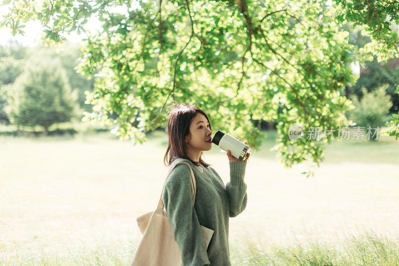 年轻的亚洲女人喝着保温瓶里的咖啡，在大自然中漫步。