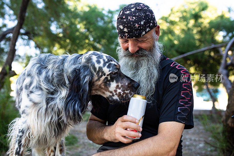 一只狗嗅着一个留着胡子、戴着大手帕的老人手里拿着的一罐饮料