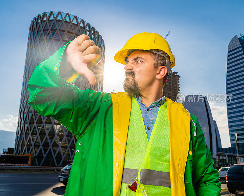年轻的工程师戴着黄色安全帽，穿着绿色防护服，在现代化的商业建筑和建筑工地前看着摄像机，并显示拇指向下的手势。镜头光晕。低角度的观点
