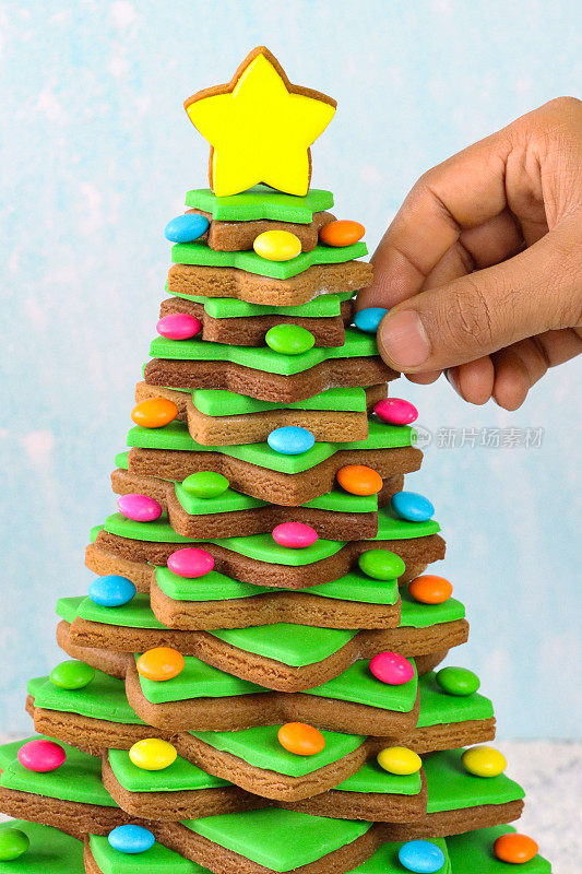 近距离拍摄，一个无法辨认的人把涂有多种颜色糖果的巧克力放在圣诞树上，圣诞树上堆叠着姜饼星饼干，上面覆有绿色方糖糖霜，背景是浅蓝色