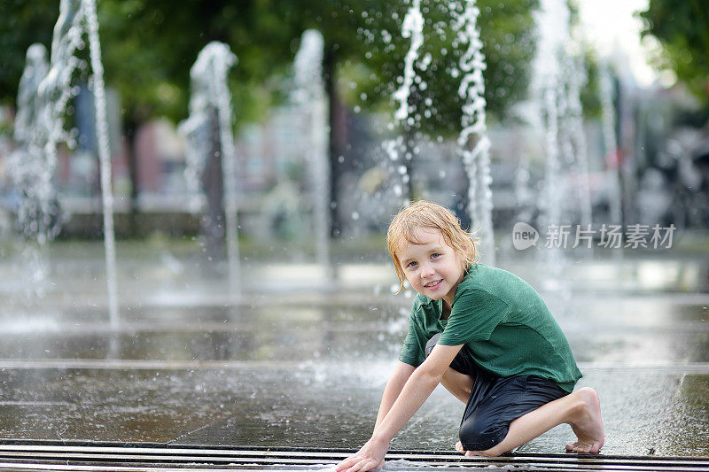一个阳光明媚的夏日，小男孩在城市喷泉的喷泉之间的广场上玩耍。城市孩子们活跃的夏季休闲活动。