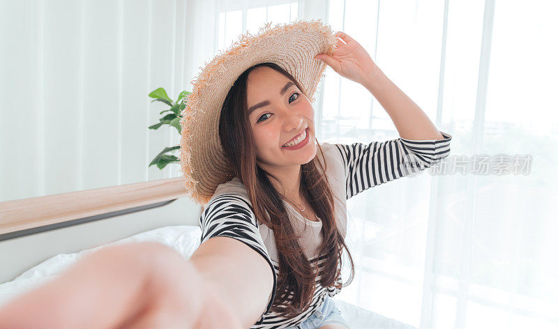 年轻的亚洲女性博主微笑在夏天的休闲服装和草帽与facetime视频通话智能手机在卧室。