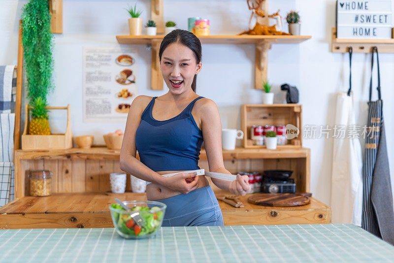 照片的年轻美丽的亚洲妇女女士穿着运动服快乐做腰测量检查她的训练，锻炼和饮食的结果。她家厨房桌上的樱桃番茄沙拉是为了减肥和健康的生活方式