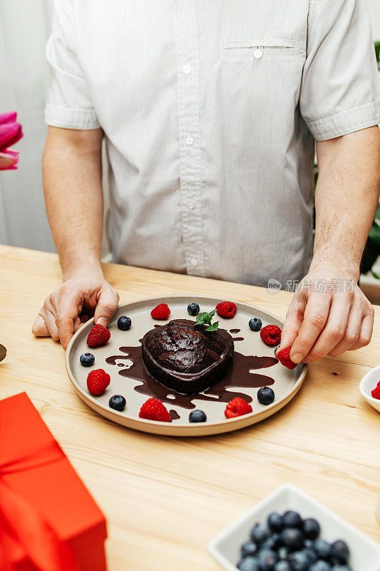 一名男子用新鲜浆果和巧克力糖衣装饰巧克力蛋糕的心形。情人节、母亲节、妇女节的礼物、惊喜