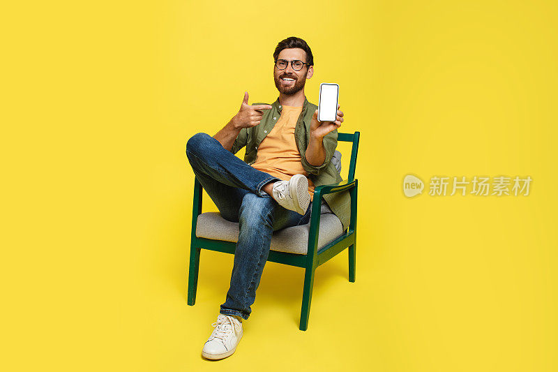 快乐的男人穿着便服指着手机空屏幕，坐在扶手椅上黄色背景