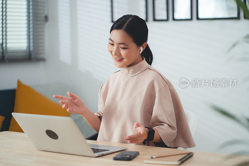 亚洲女性戴着耳机，喜欢坐在家里的办公室里，用笔记本电脑在线工作，进行视频会议。