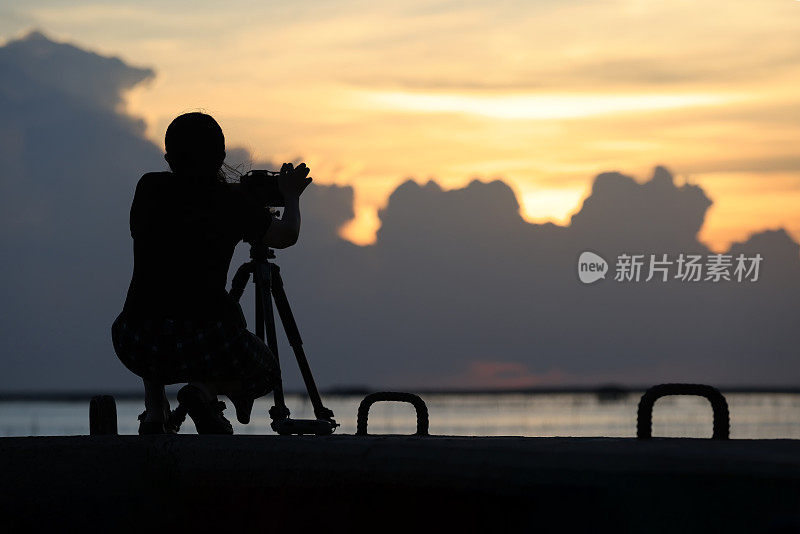 一个女人在夕阳下拍照的剪影。摄影师用剪影在桥上拍摄日落照片。准备好相机和三脚架，准备在桥上拍一张日落照片。