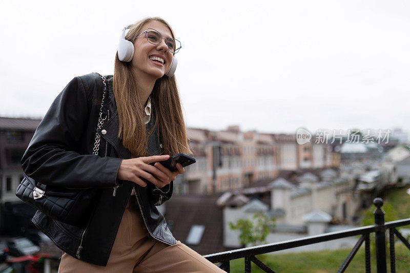 一个年轻的学生戴着耳机，手里拿着手机，在城市风景的背景下听着悦耳的音乐