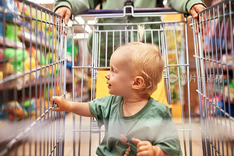 超市里可爱的宝宝被妈妈推着购物车的特写。妈妈和金发碧眼的小女儿穿着相似的绿色衣服逛街，玩得很开心。合家周末，快乐童年。