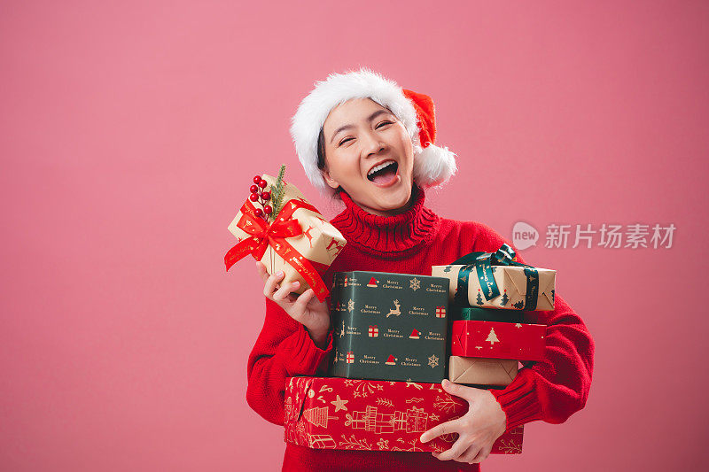 亚洲女人戴着圣诞老人的帽子，开心地微笑着，手里拿着许多礼盒，孤立在粉红色的背景上。