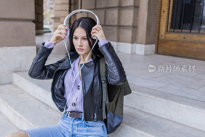 年轻漂亮的女大学生背着双肩包坐在大学楼梯上。她戴着耳机，穿着皮夹克，等待着大学课程的开始。