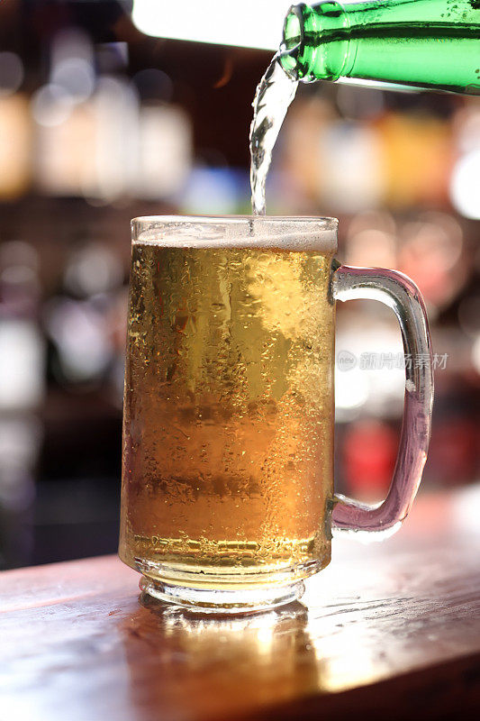 啤酒从绿色啤酒瓶倒进玻璃大酒杯的特写图片，啤酒杯上覆盖着凝结的气泡，在啤酒上形成泡沫的泡沫头，木制吧台，酒吧背景，焦点在前景上