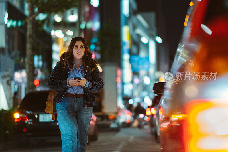 年轻女子拿着智能手机，晚上在城市里行走